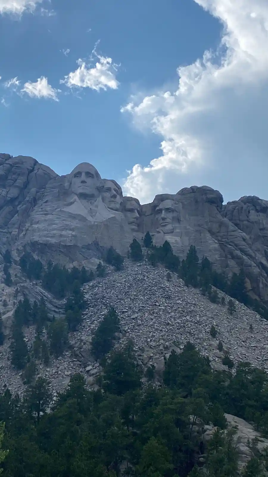 A photo of Mount Rushmore showing the carved faces of four U.S. presidents against a blue sky with clouds, with evergreen…