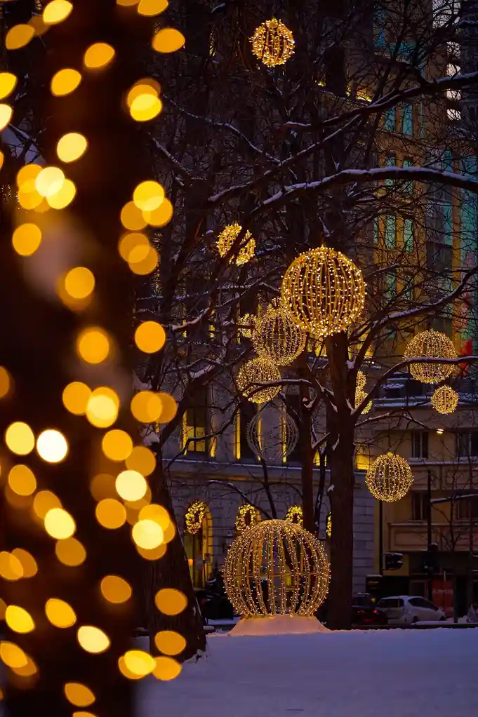 A snowy winter night scene with bare trees decorated with large, glowing golden wireframe spheres and strings of lights…