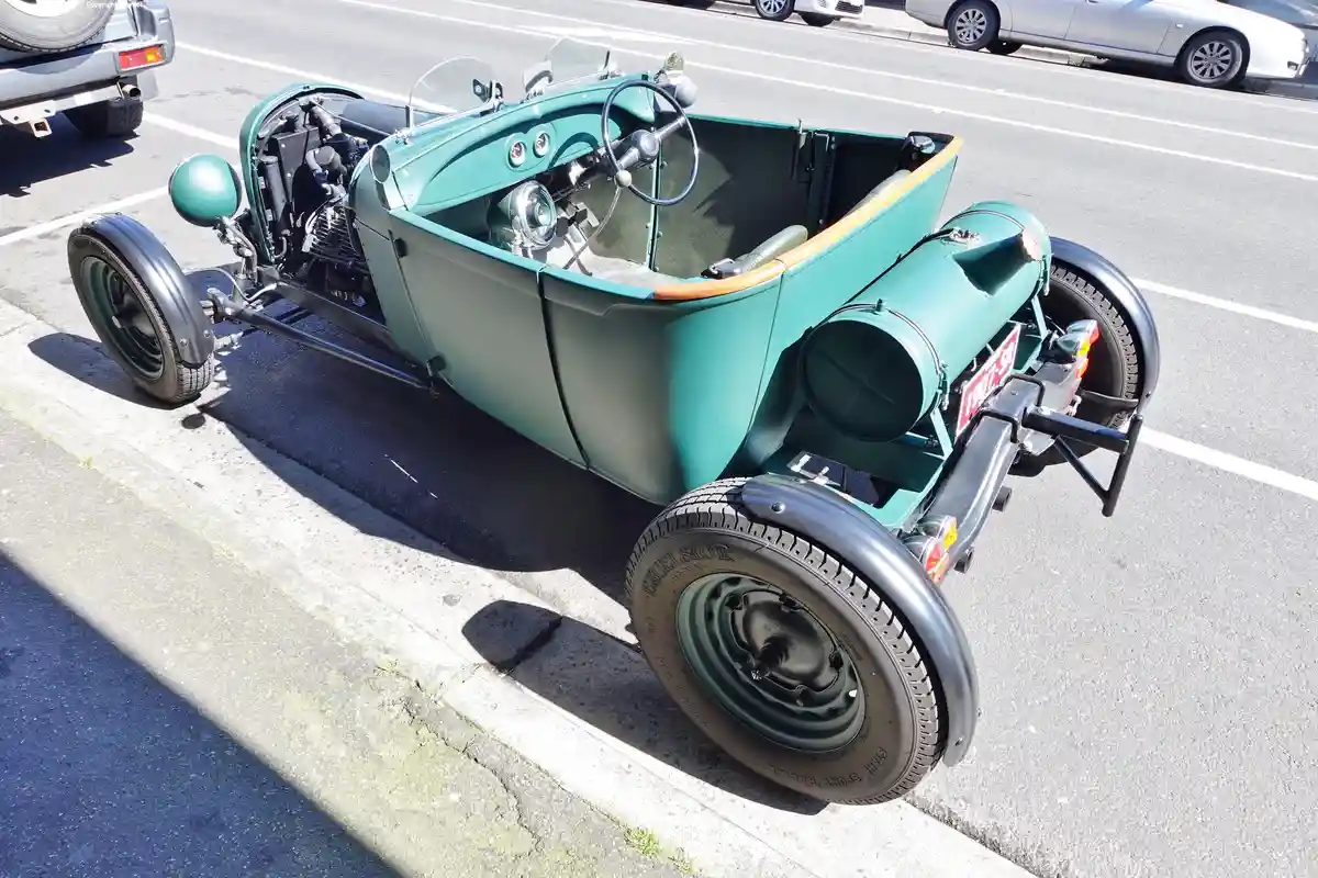 A teal vintage open-top car with exposed engine and large fuel tank is parked on a street, its license plate reads…