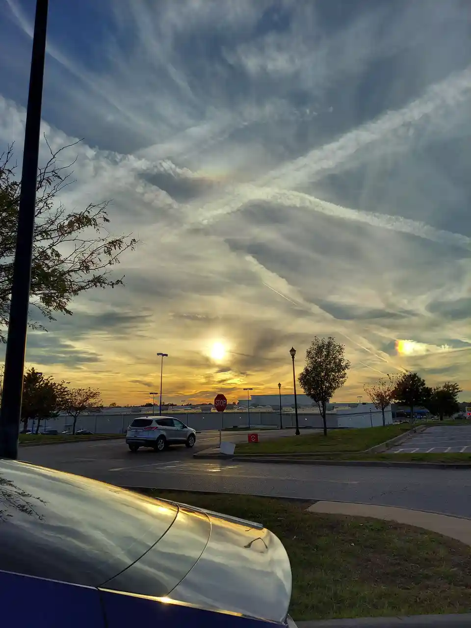 A parking lot at sunset with a silver SUV driving away, a stop sign visible, and a dramatic sky filled with streaky clouds…