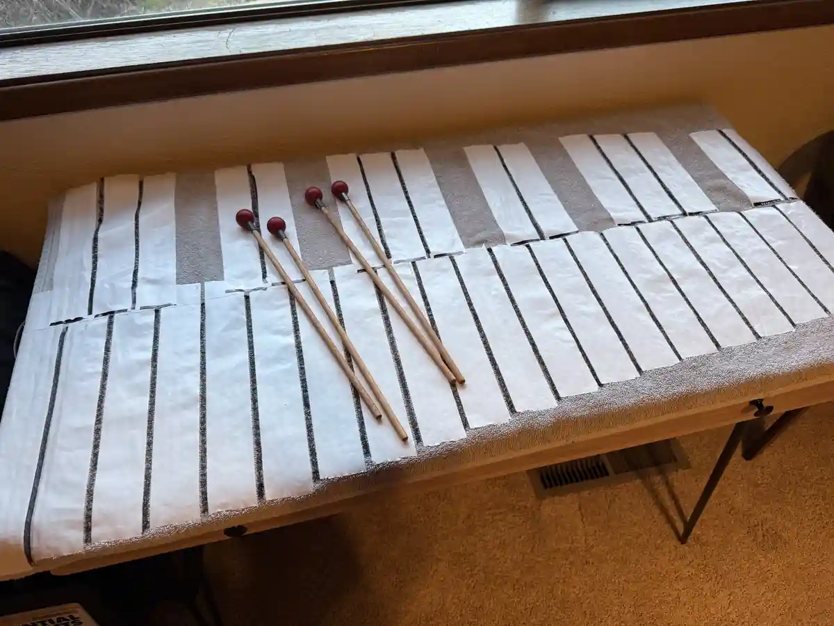 A wooden table covered with a towel, on which white toilet paper rolls are arranged in rows to mimic piano keys, with four…
