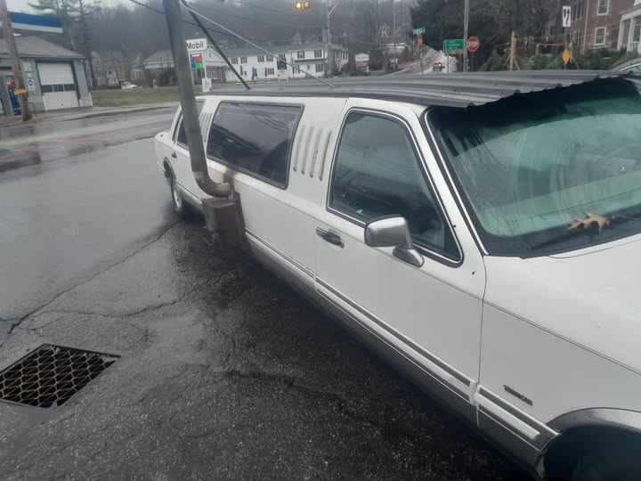 A white stretch limousine is parked on a wet street, visibly damaged with its roof torn off and hanging down. A utility pole…