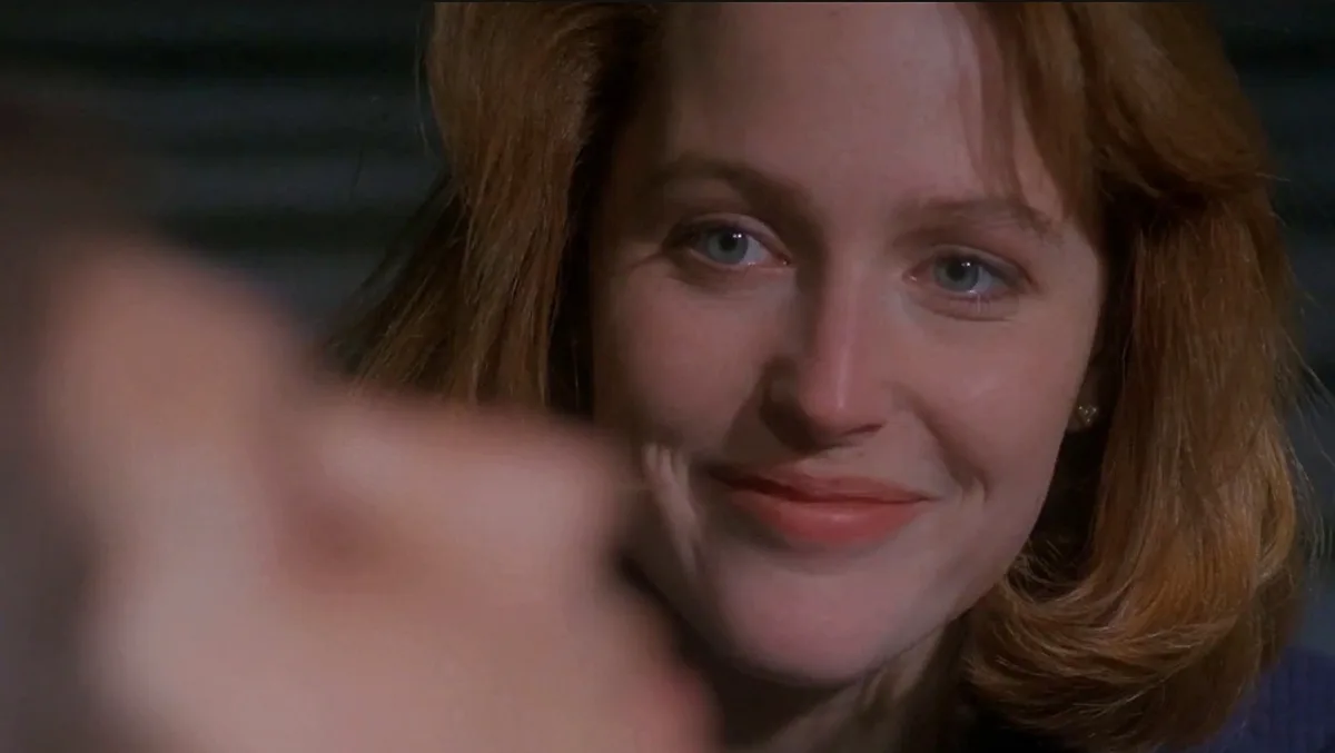 a close-up of a woman with reddish-brown hair and blue eyes smiling faintly while looking at a blurred hand in the…