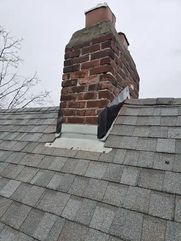 A low-angle photo shows a brick chimney rising from a gray asphalt shingle roof under an overcast sky, with bare tree…