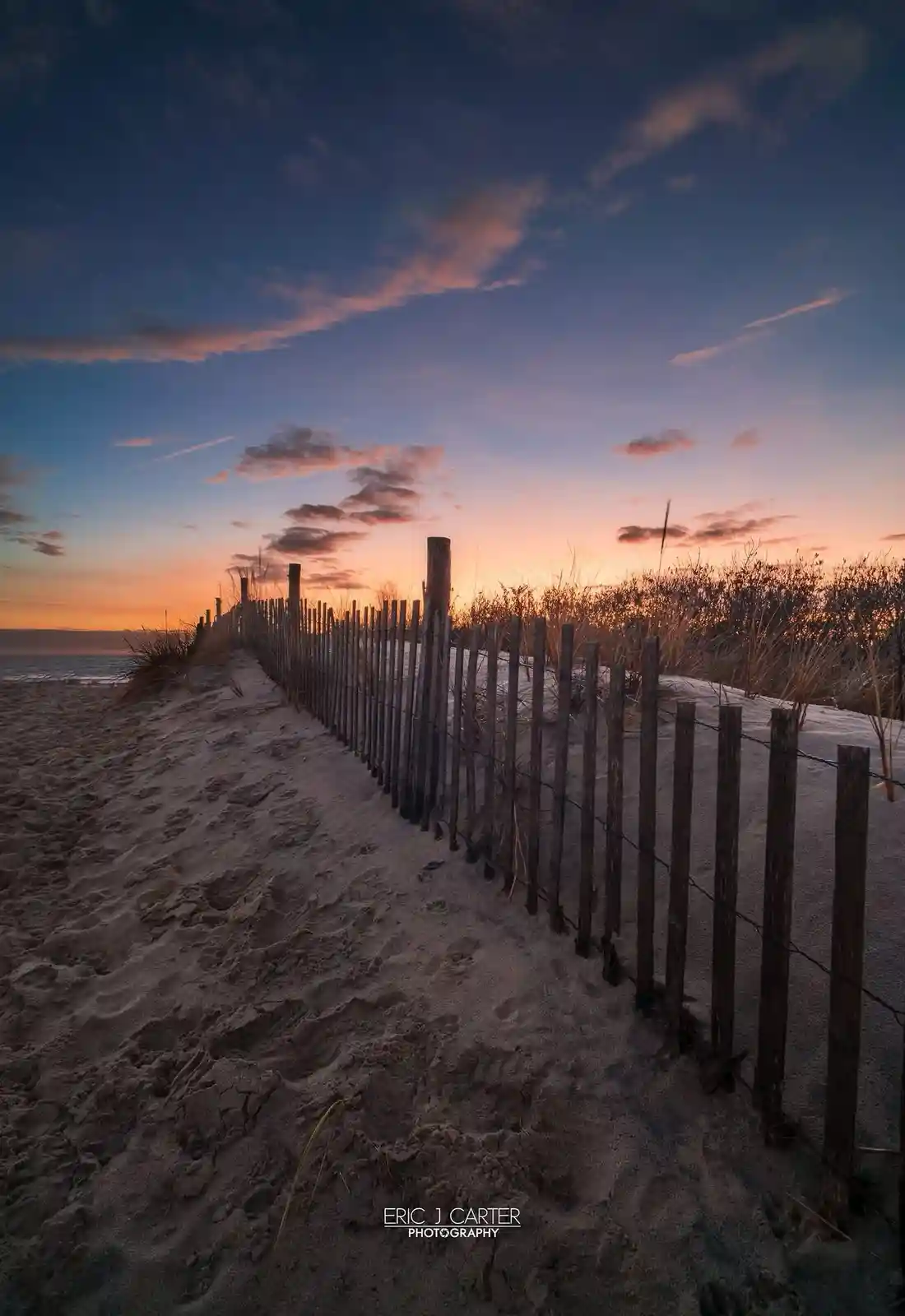 A serene beach scene at sunset, featuring a wooden fence running along a sandy dune path, with the ocean visible in the…
