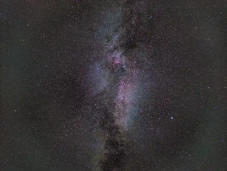 A vertical photograph of the Milky Way galaxy, showing a dense band of stars, cosmic dust, and colorful nebulae stretching…