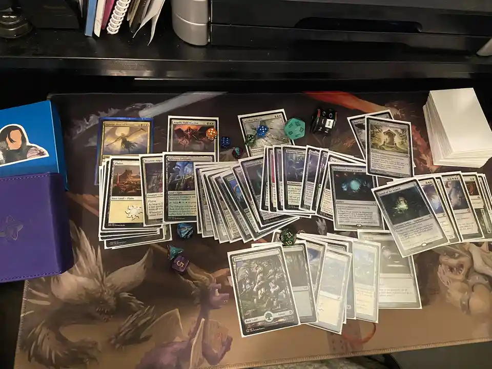An overhead shot of a cluttered Magic: The Gathering playmat covered in scattered cards, dice, and a stack of blank papers…