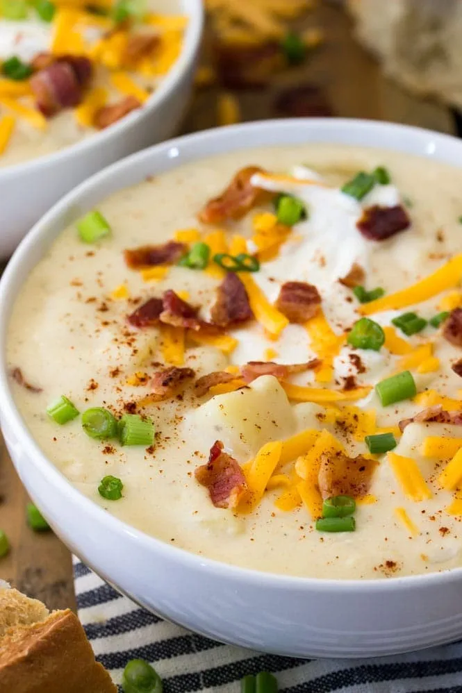 A close-up photo of two white bowls filled with creamy potato soup, topped with crispy bacon bits, shredded cheddar cheese…