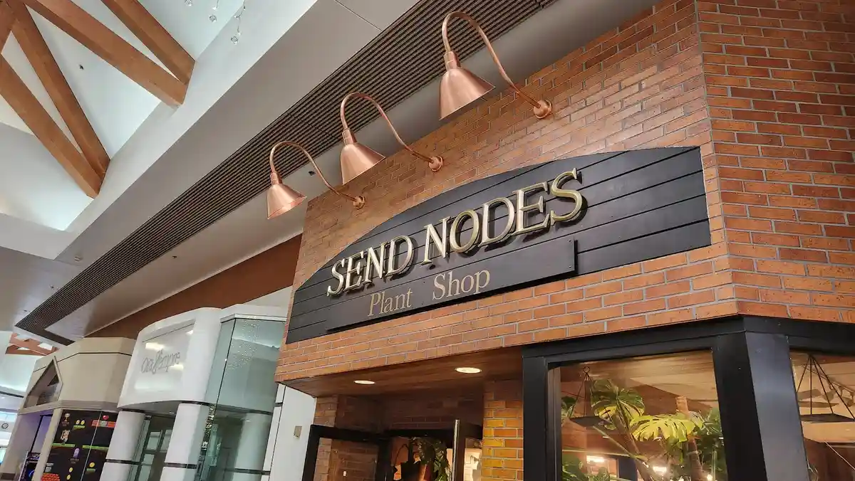 A storefront with a black sign reading 'SEND NODES Plant Shop' mounted on a brick wall, illuminated by three copper-colored…