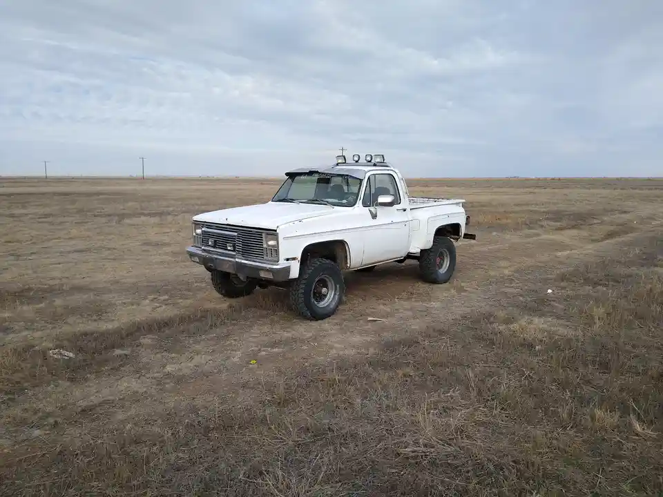 A white, lifted, older model pickup truck with oversized tires and roof-mounted lights is parked alone in a vast, dry…
