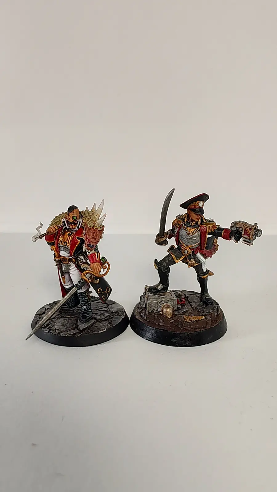 Two intricately painted fantasy miniatures stand side-by-side on a plain white surface. The figure on the left, a heavily…