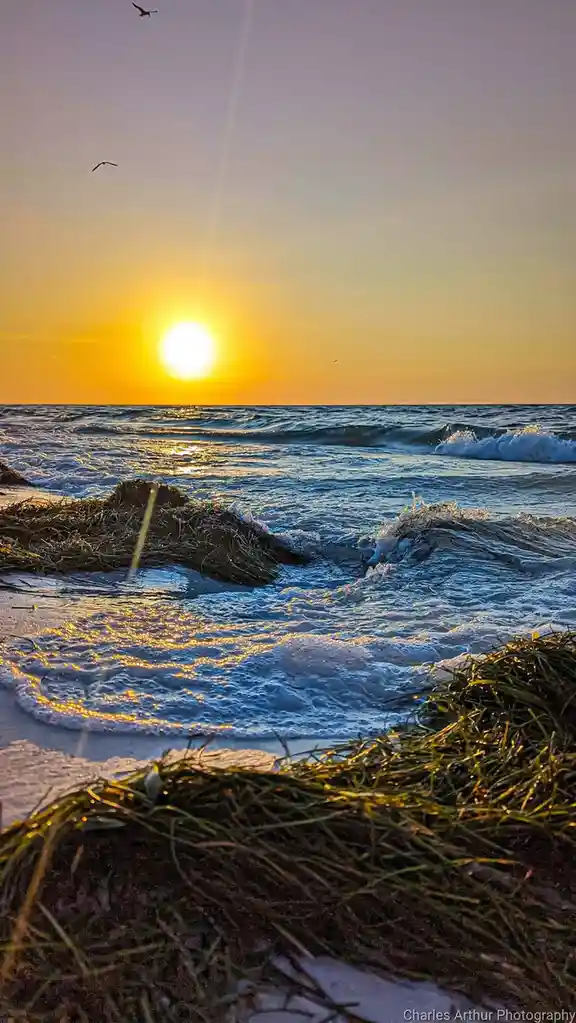 A serene beach sunset photo showing the sun low on the horizon over the ocean, with waves crashing onto the shore and…