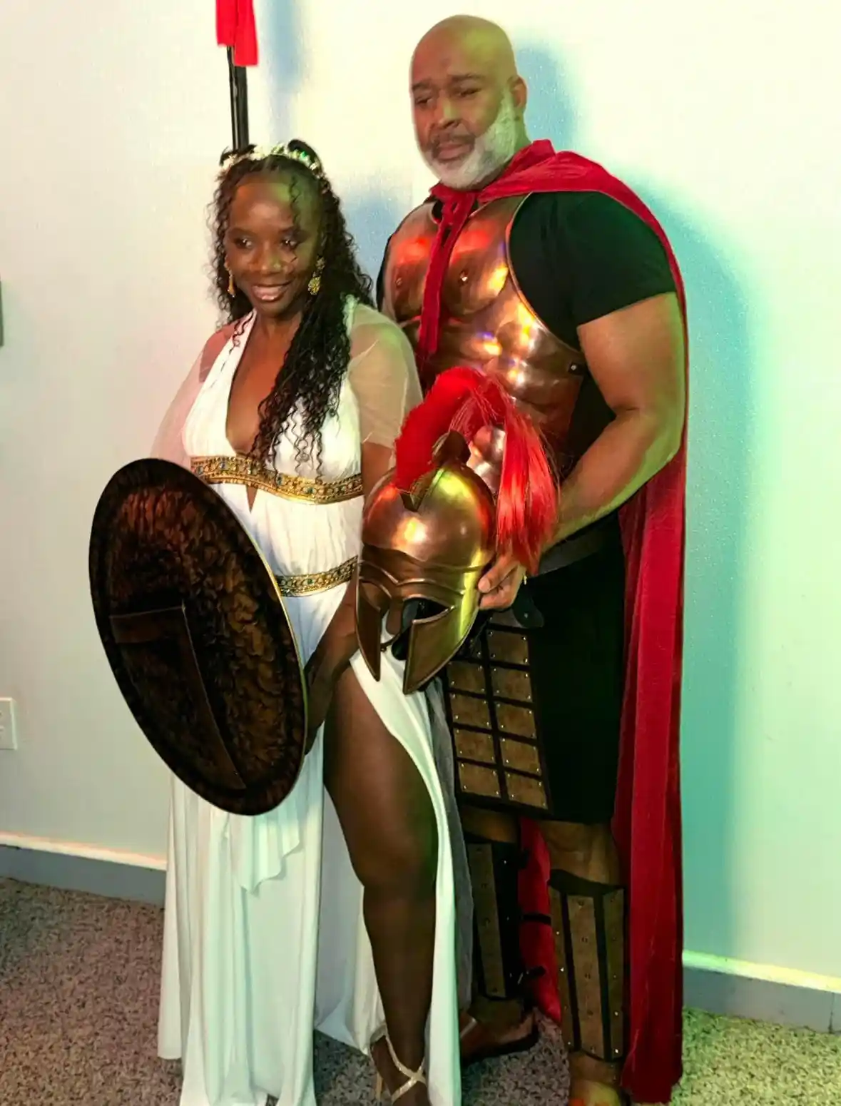 A man and woman dressed in ancient Greek warrior costumes pose indoors. The woman wears a white toga-style dress with a gold…