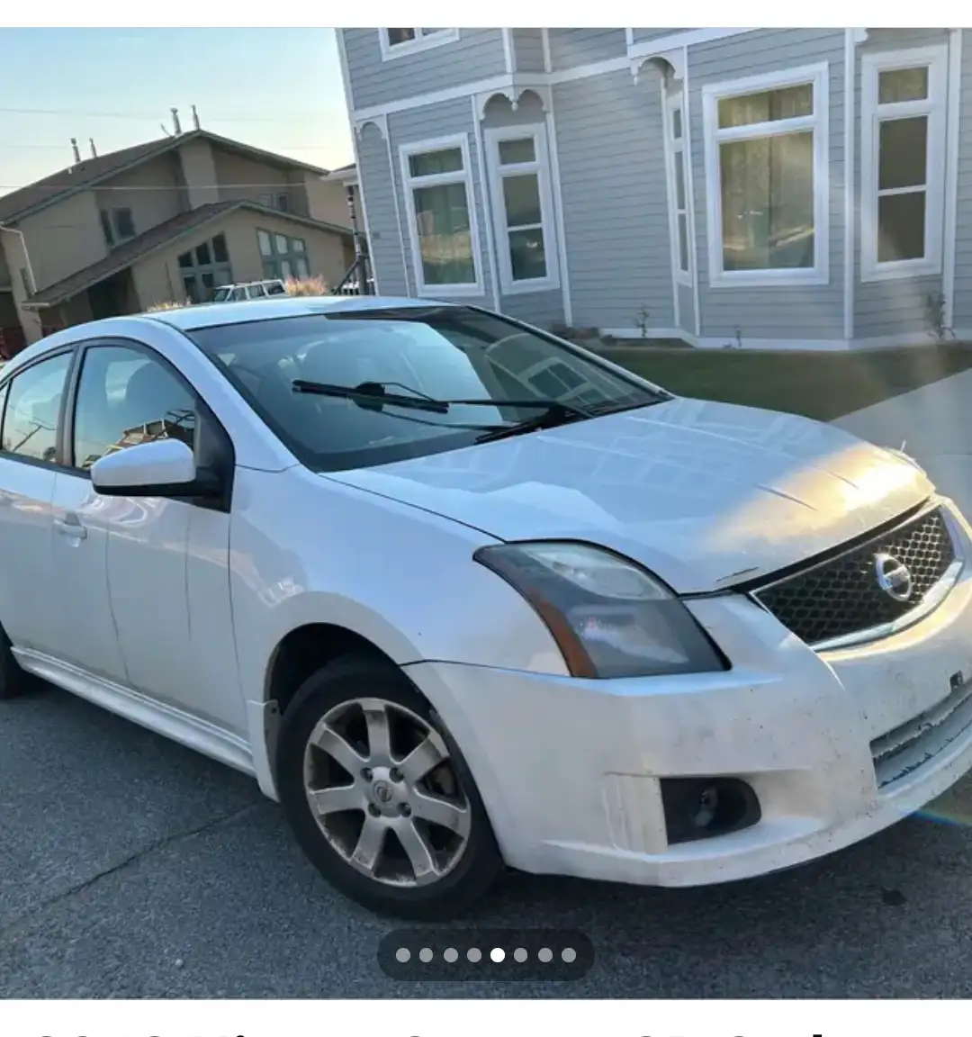 A white 2006 Nissan Sentra is parked in front of a light gray house with white trim. The car's front bumper and hood show…
