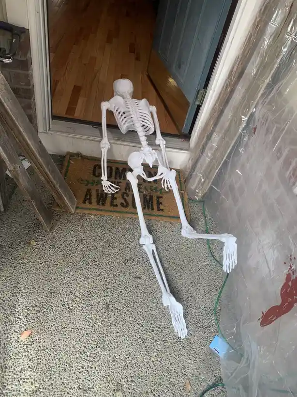 A plastic skeleton decoration is lying on its back on a concrete porch, positioned in front of an open door with a doormat…