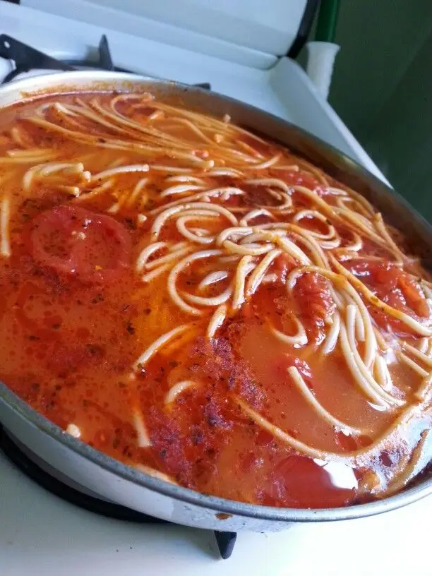 A close-up photo of a pan on a stovetop filled with spaghetti noodles submerged in a red tomato sauce, with visible tomato…