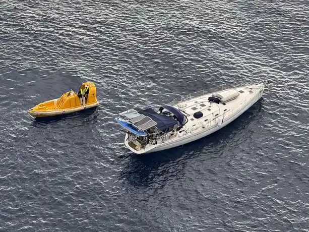 An aerial view shows a white sailboat with solar panels on its deck floating in choppy dark blue water, alongside a bright…