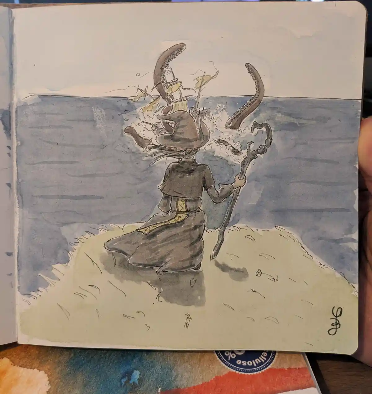 A watercolor sketch shows a witch in a pointed hat and dark robe standing on a beach, facing the ocean. Behind her, a large…