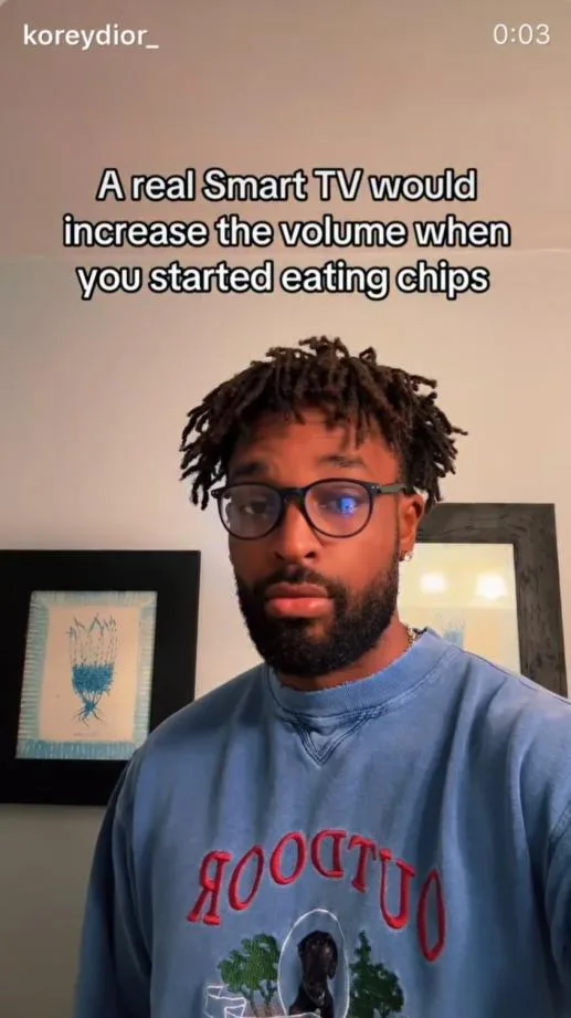 A man with dreadlocks and glasses stares intently at the camera while wearing a blue sweatshirt with 'OUTDOOR' written on…