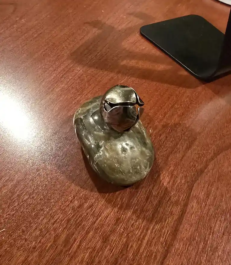 A small, metallic duck figurine with a bell-shaped head sits on a wooden table, its body appearing worn and textured. The…
