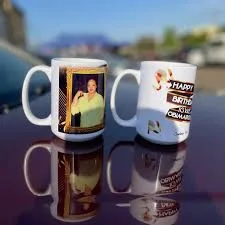 Two white ceramic mugs sit on a reflective dark surface outdoors. The mug on the left features a photo of a smiling man in a…