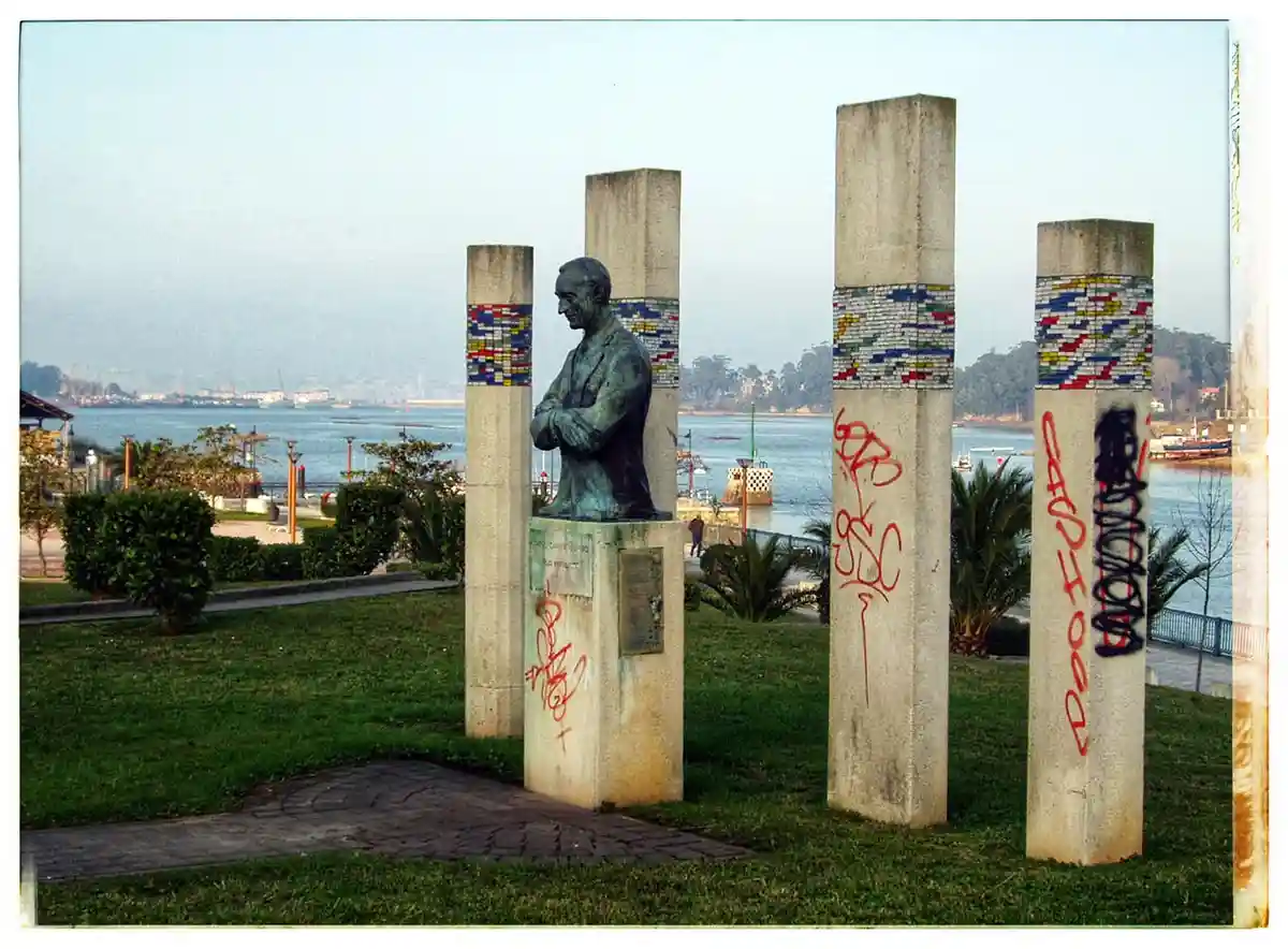 A bronze bust statue of a man with arms crossed stands on a pedestal among four tall concrete pillars, all of which are…