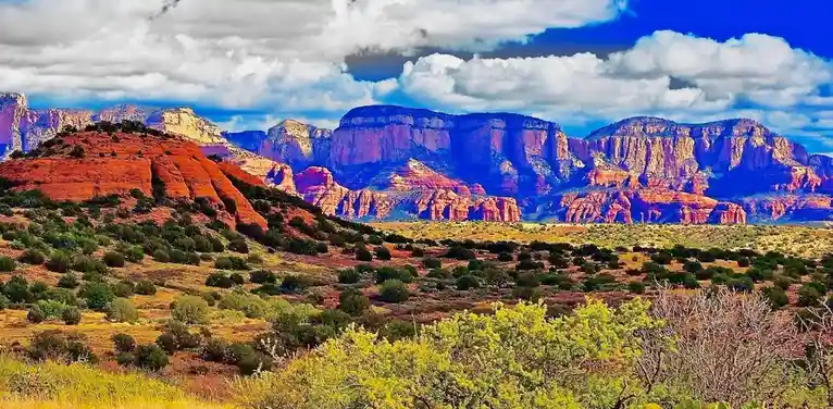 A vibrant, heavily saturated landscape photo of red rock formations and desert scrub under a dramatic blue sky with white…