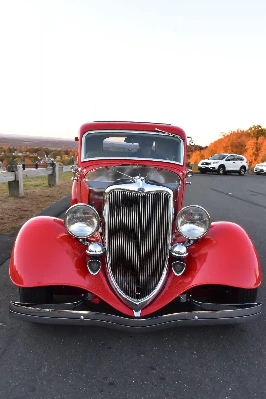 A shiny red vintage Ford truck is parked in a scenic autumn overlook, with a man in sunglasses visible in the driver's seat…