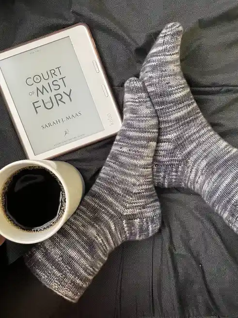 An overhead shot shows a person's feet in gray and white striped socks resting on dark gray bedding, next to a white mug of…