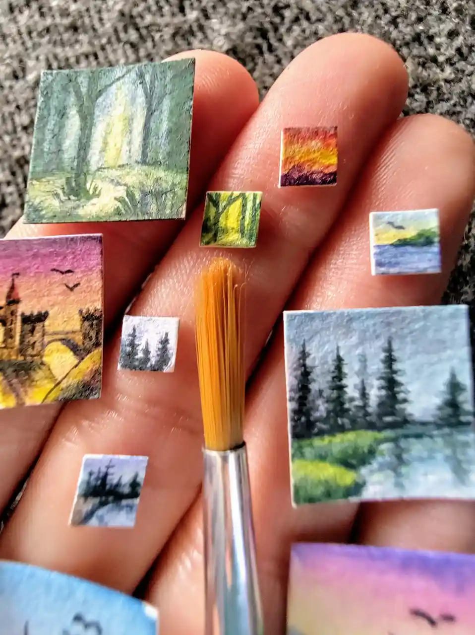 A hand holds numerous tiny square paintings depicting various landscapes like forests, sunsets, and castles, alongside a…
