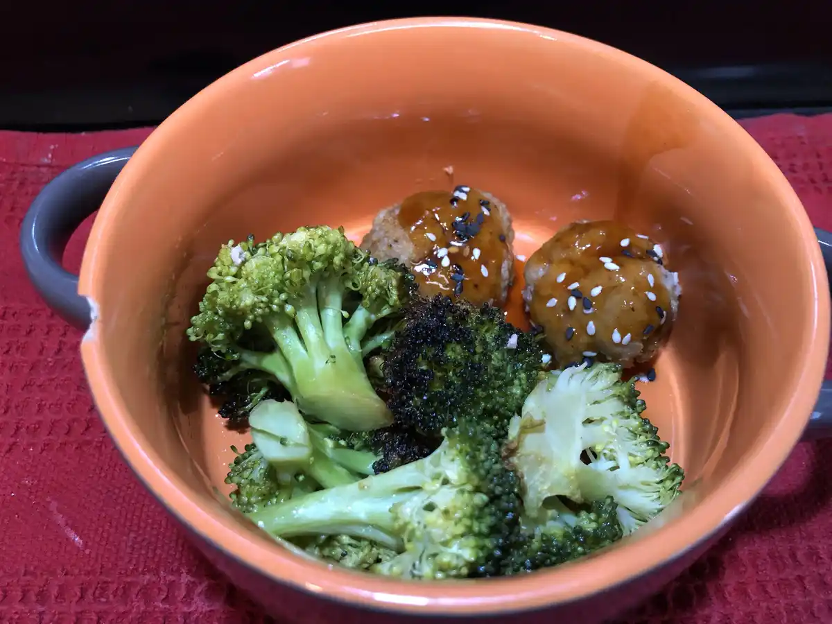 An orange ceramic bowl with grey handles contains two meatballs covered in brown sauce and sprinkled with black and white…