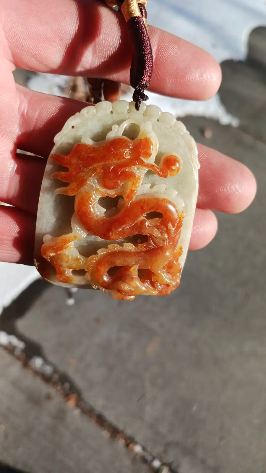 a hand holds a carved jade pendant featuring a stylized dragon with orange-red and white coloring, suspended from a braided…
