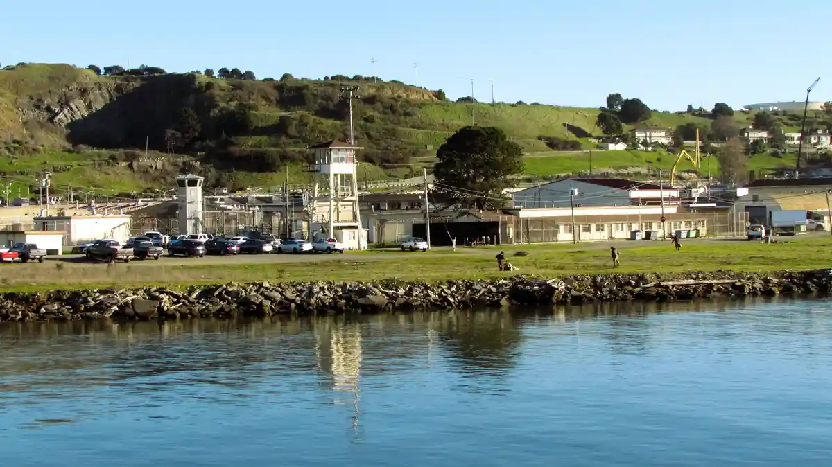 A wide shot of a prison complex with watchtowers, fences, and parked cars, viewed from across a calm body of water. The…