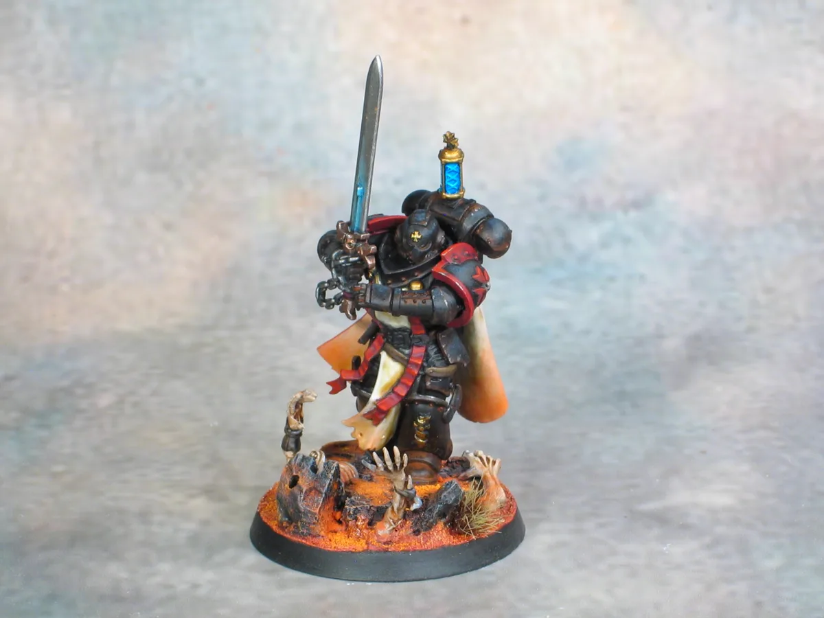 A painted miniature of a heavily armored warrior, likely from Warhammer 40,000, stands on a base with orange terrain and…