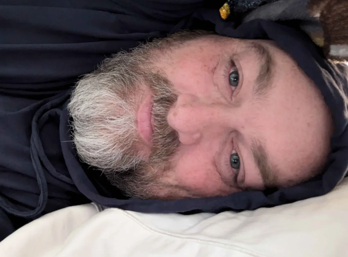 A man with gray hair and a beard lies on his back, looking up at the camera with a tired expression, wearing a dark hoodie…