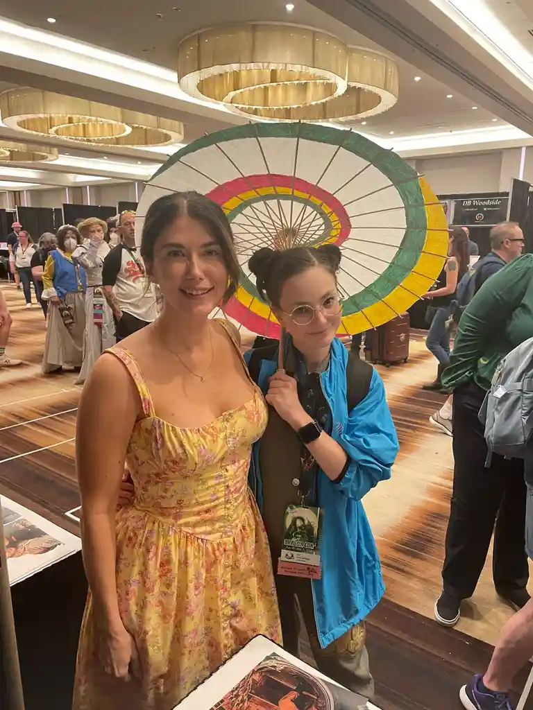 Two women pose for a photo at a convention hall; one wears a yellow floral dress and smiles, while the other, in a blue…