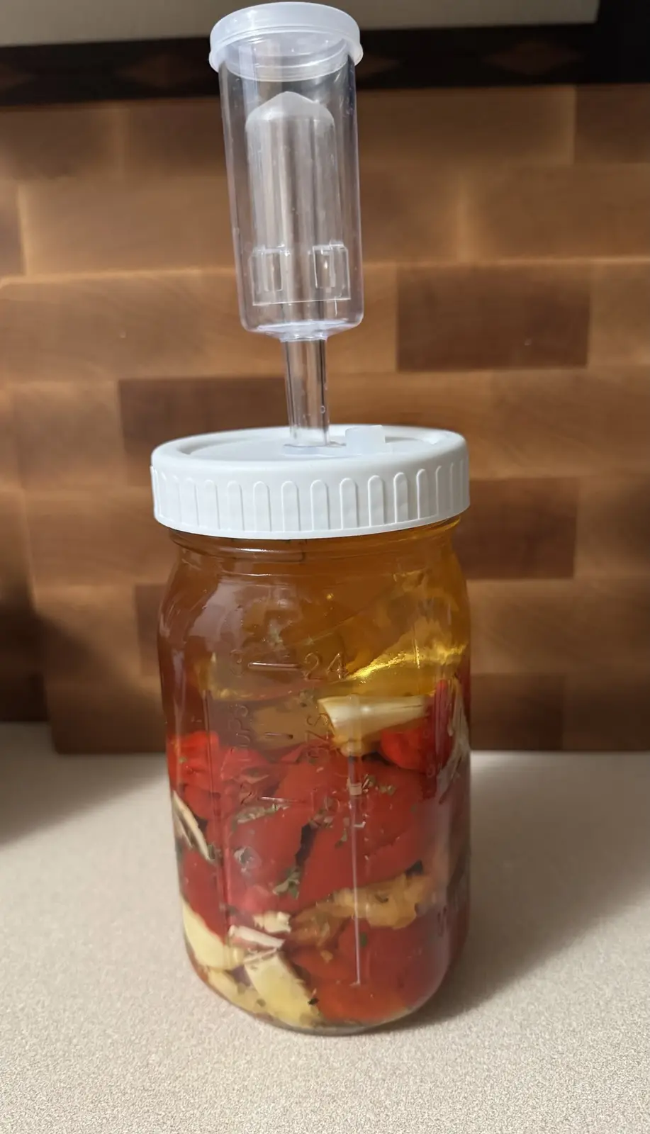 A glass mason jar filled with chopped red peppers, garlic cloves, and liquid, sealed with a white lid and a clear airlock…