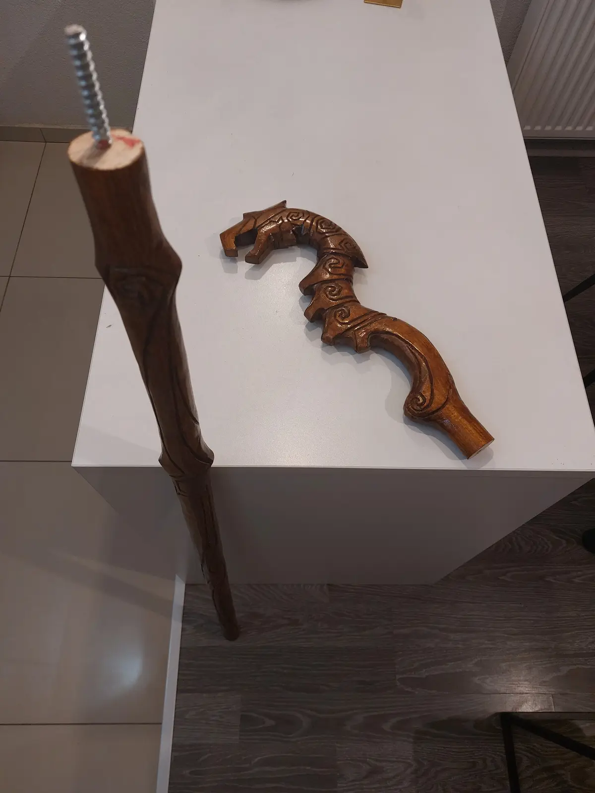 A wooden walking cane with an ornate, carved handle lies on a white display table, while its shaft has a large metal screw…