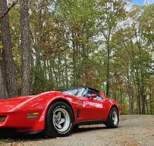 A shiny red classic Chevrolet Corvette sports car is parked on a gravel road surrounded by tall trees with autumn foliage…