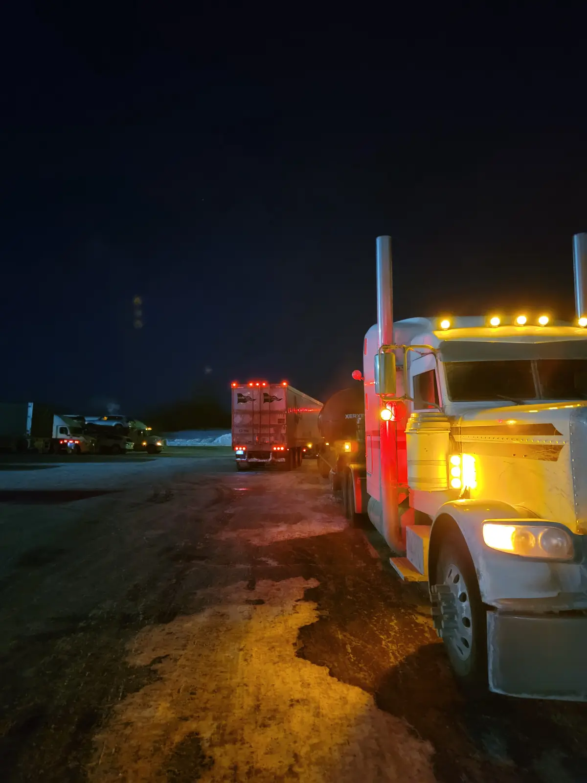 A nighttime photo shows a white semi-truck with its headlights and roof lights on, parked in a snowy lot with other trucks…
