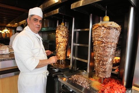 A chef in a white uniform and hat stands beside two large vertical rotisserie spits of meat, using a long knife to shave…