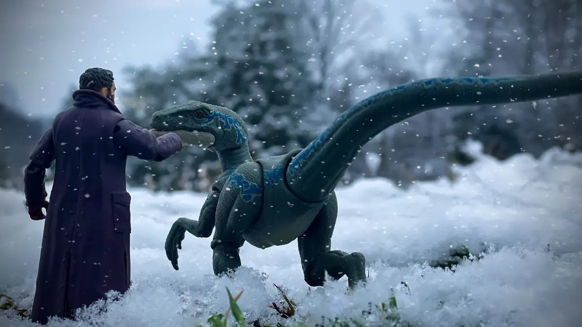 A person in a long purple coat stands in a snowy outdoor scene, gently touching the snout of a blue and grey velociraptor…