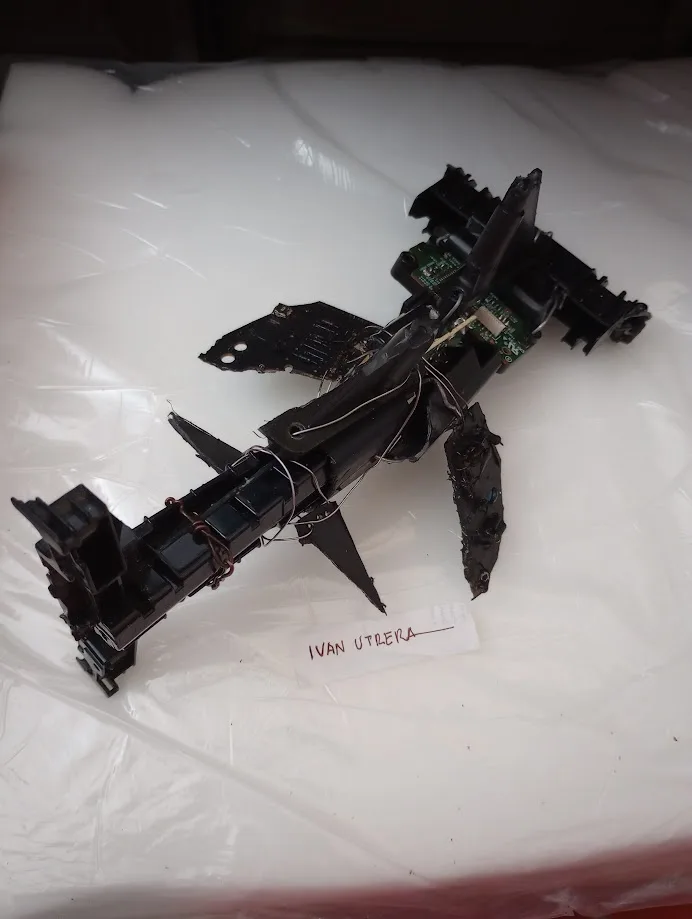 A disassembled black electronic device, possibly a drone or remote-controlled vehicle, lies on a white plastic surface with…
