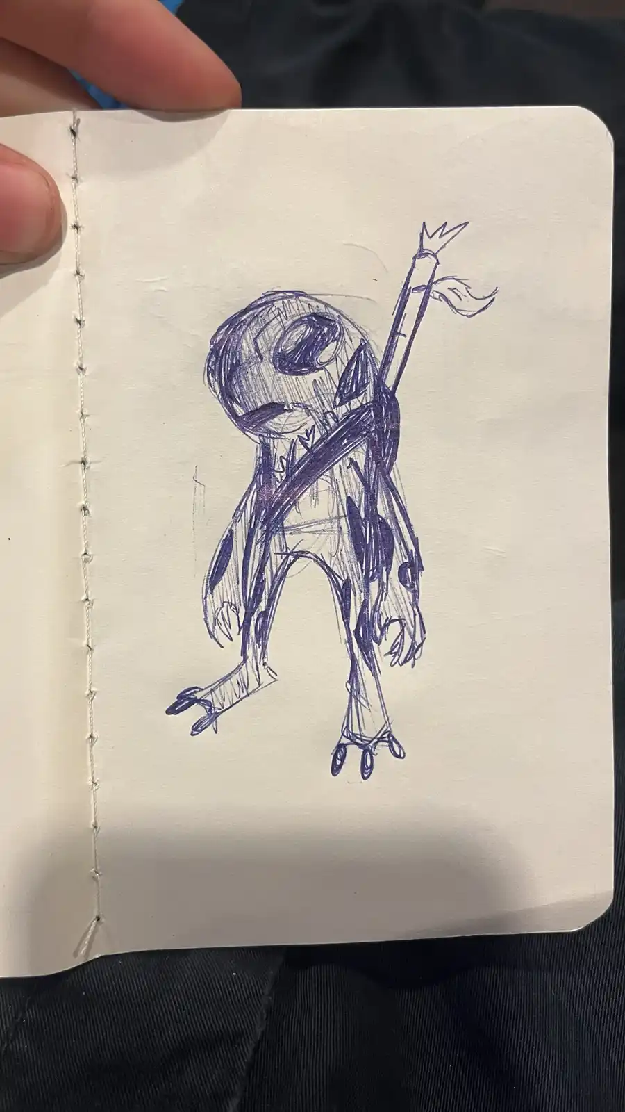 A hand holds open a stitched notebook displaying a crude blue ink drawing of Jack Skellington, the skeletal character from…