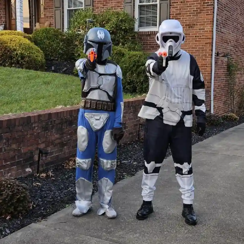 Two people in Star Wars costumes stand on a sidewalk in front of a brick house, both pointing orange-tipped toy blasters at…