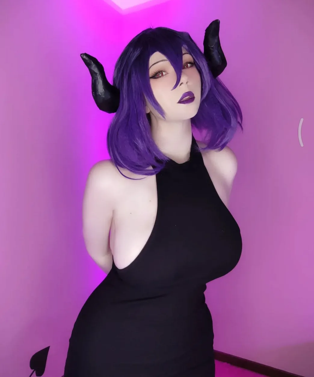 A person with purple hair and black horns poses in a black dress against a pink-lit wall, with the overlaid text reading…