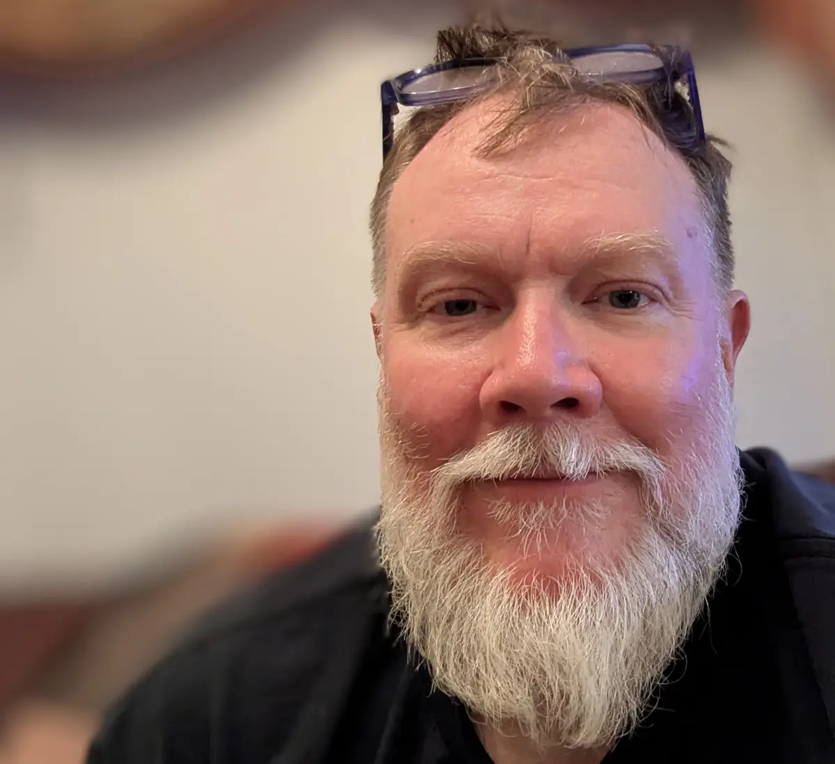 A close-up photo of a middle-aged man with a white beard and glasses perched on his head, looking directly at the camera…