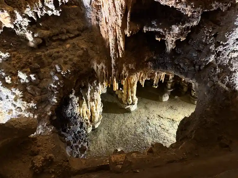 A dimly lit cave interior with rugged, textured rock walls and ceilings, featuring prominent stalactites hanging down and…