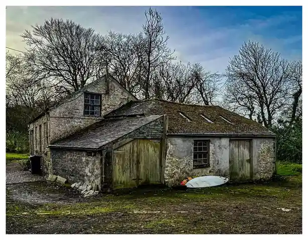 A weathered, dilapidated stone building with a shingled roof and wooden doors sits in a grassy, muddy yard. Bare trees frame…
