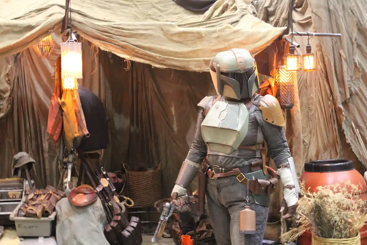 A person in a Mandalorian armor costume stands in a rustic, tent-like setting with hanging lanterns and various props like…
